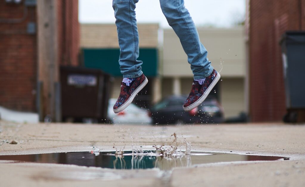 A young person in May jumping over a puddle, only showing their shoes above the puddle. 